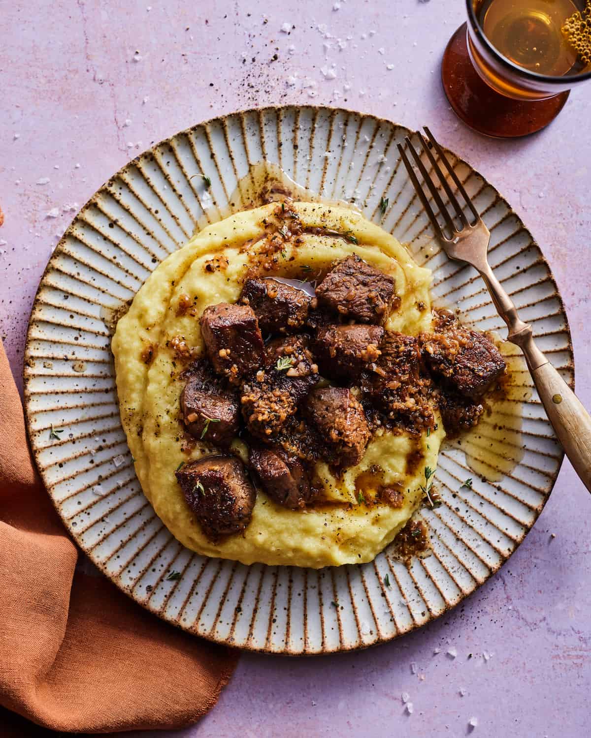 An overhead shot of a ceramic fluted plate with garlic butter steak bites over creamy polenta. There is a fork resting on the right rim of the plate and a burn orange linen napkin to the left side of the plate. An amber glass of sparkling water is at the top right.