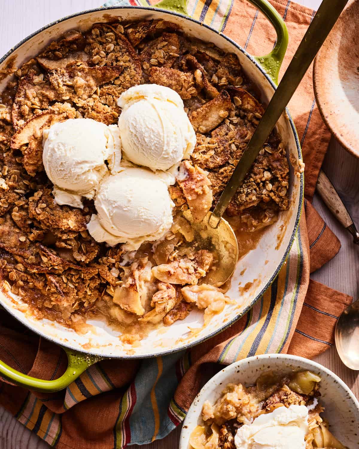 An overhead shot of apple crisp in a green braiser with three scoops of vanilla ice cream on top and a gold serving spoon resting in the bottom right of the braiser. The braiser is on a orange blue and green striped kitchen towel and there is a speckled ceramic bowl of apple crisp and ice cream in the bottom right corner.