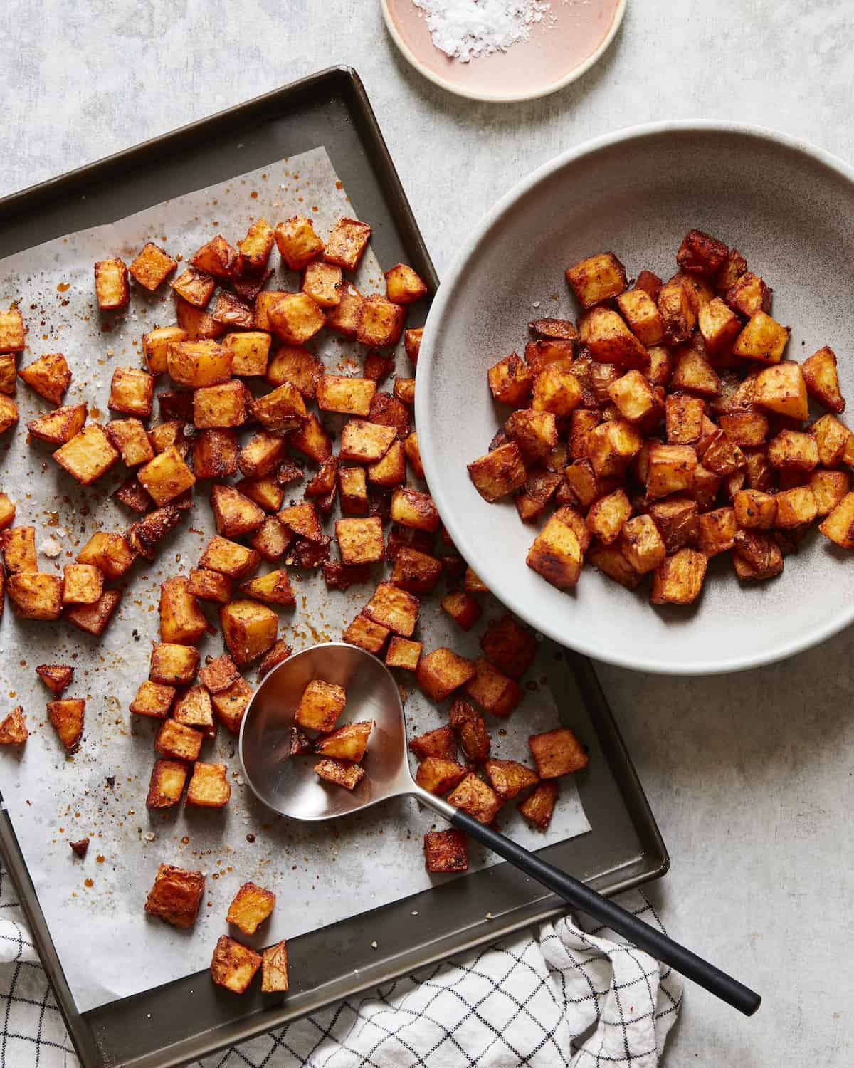 A parchment paper-lined baking sheet with cubed roasted potatoes and a serving spoon, and a grey bowl on the side with more potatoes in it.
