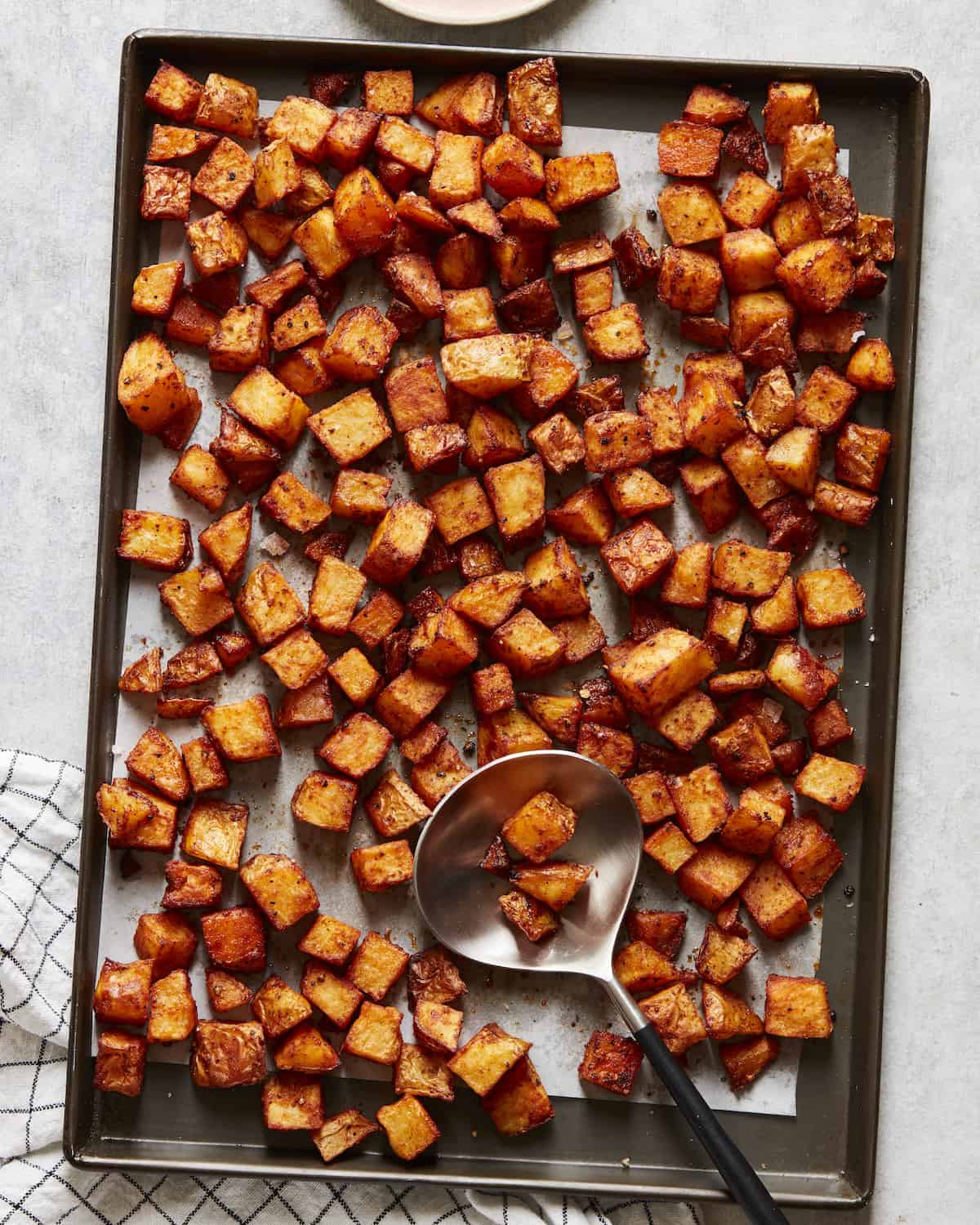 A parchment paper-lined baking sheet with cubed roasted potatoes and a serving spoon placed on a white kitchen towel.