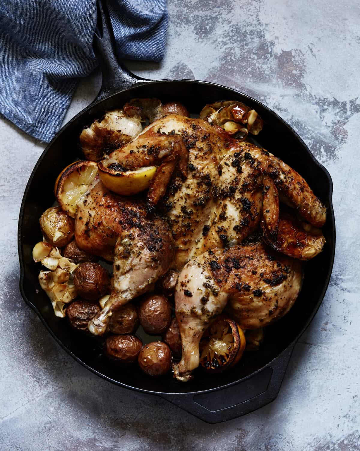 A cast iron skillet placed on a blue kitchen towel with a spatchcocked roasted chicken in it, sitting on a bed of lemon halves and baby potatoes.
