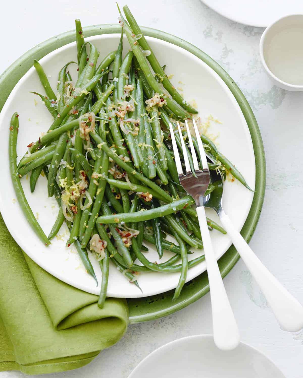 Spicy garlic green beans on a white plate with a serving fork and spoon. Beneath the white plate is a green platter and a green linen napkin on a white countertop with a white plate in the bottom right corner and a ceramic drinking glass and plate in the top right corner. 