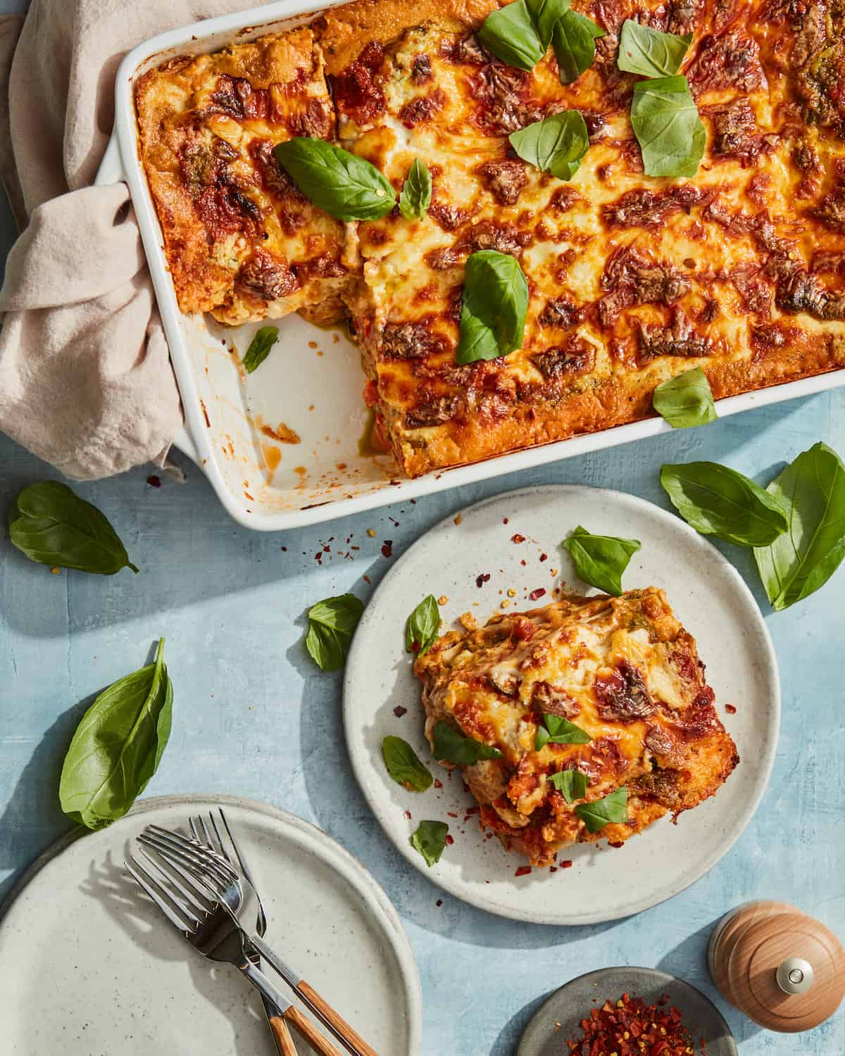 A white rectangular baking dish with lasagna garnished with basil leaves, and a corner piece cut out and served in a small ceramic plate on the side.