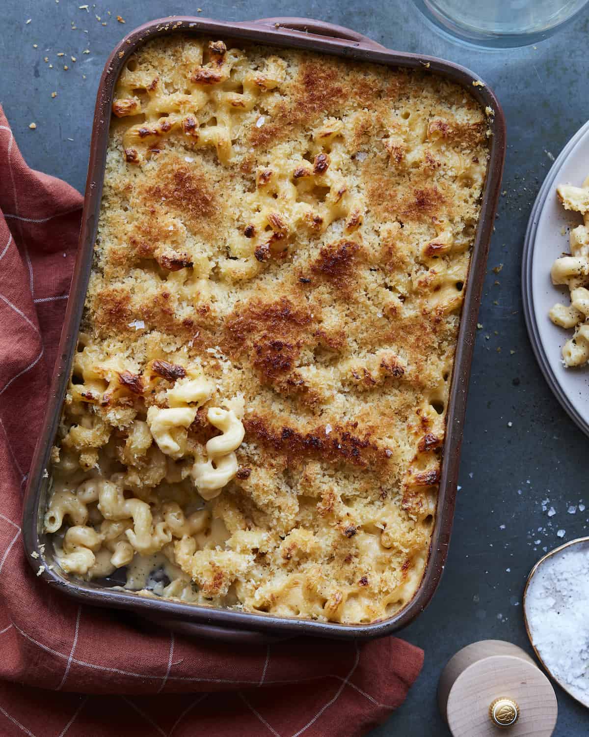 A baking dish with baked mac and cheese with a little bowl of salt and a plate with mac and cheese in the corner.