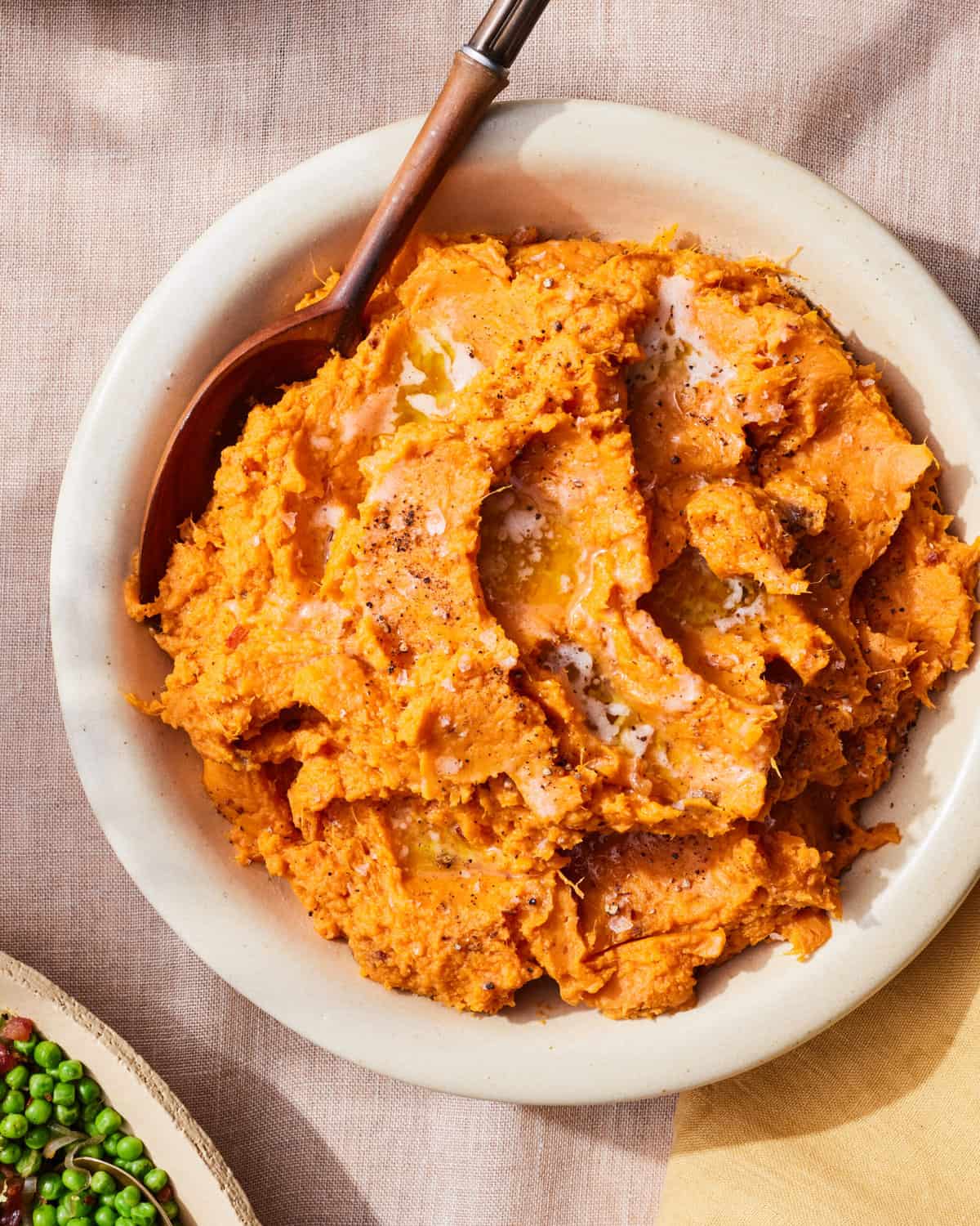 A white bowl with chipotle mashed sweet potatoes, with a bowl of green peas on the corner.