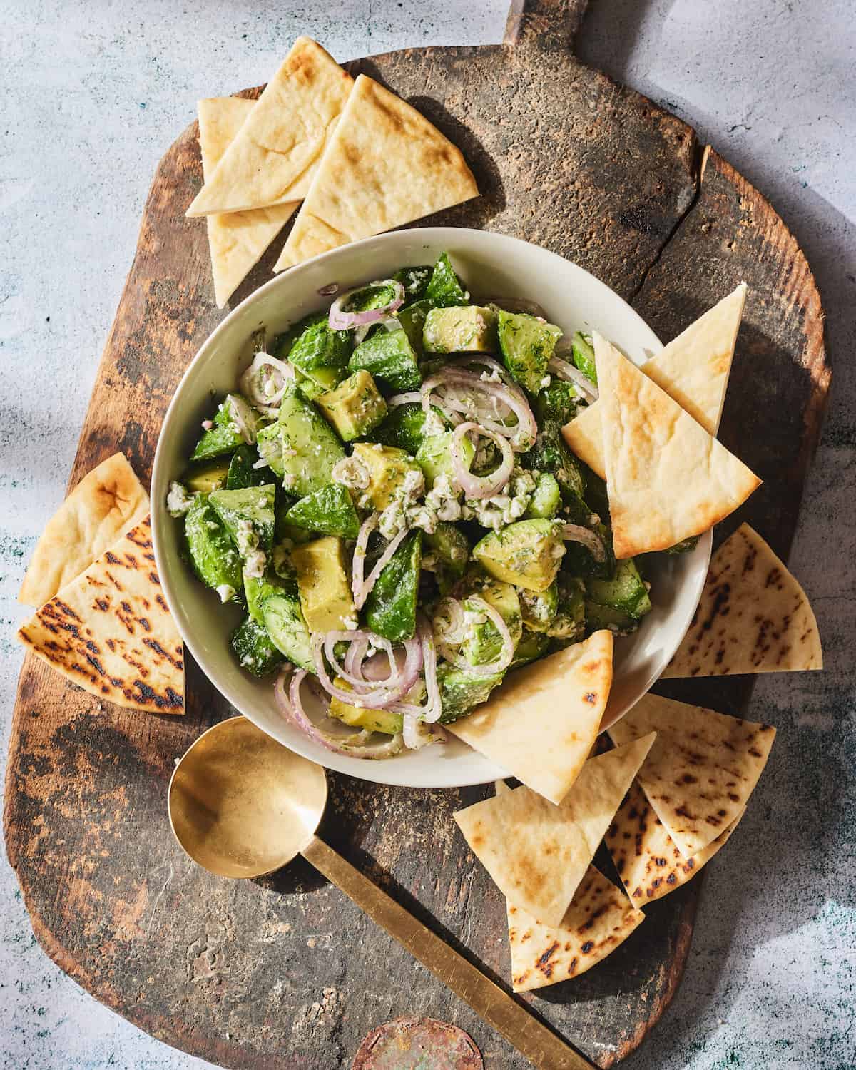 A wooden board with a white bowl of cucumber avocado feta salad garnished with sliced shallots, with pita chips and a gold spoon on the side.