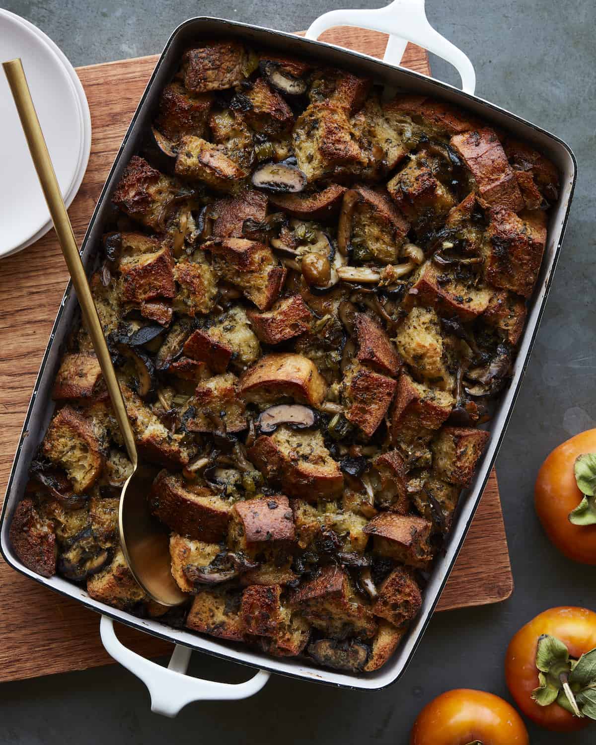 A baking dish with a gold serving spoon with mushroom stuffing, with persimmons on the side.