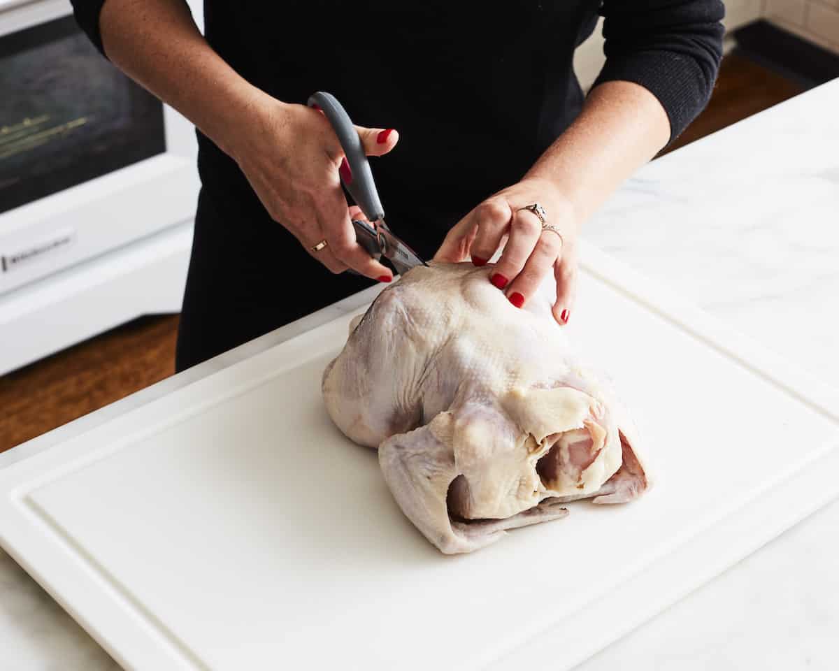 A woman cutting along one side of the backbone of a chicken with kitchen scissors, placed on a white cutting board.