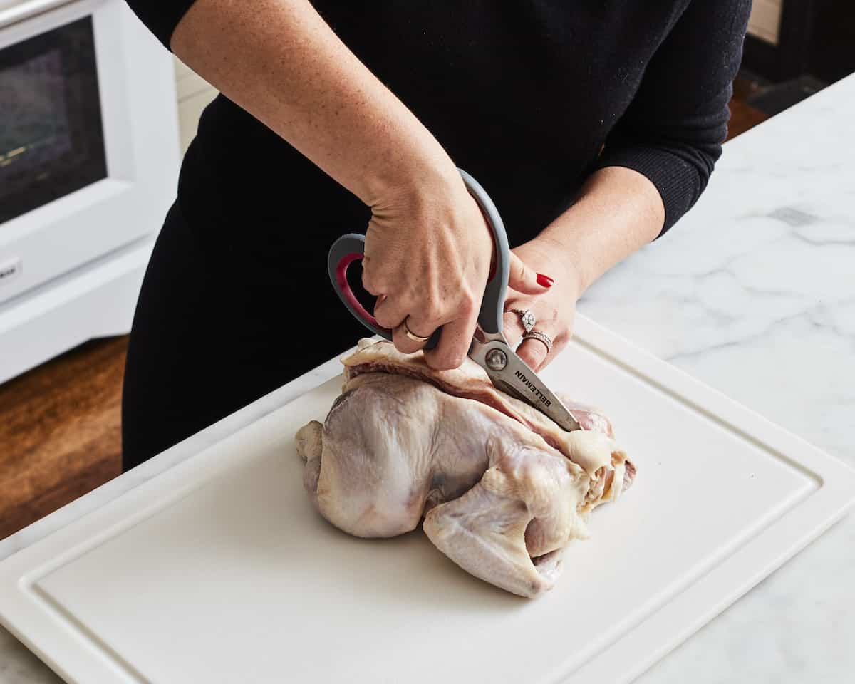 A woman cutting along one side of the backbone of a chicken with kitchen scissors, placed on a white cutting board, and the other side is already cut.