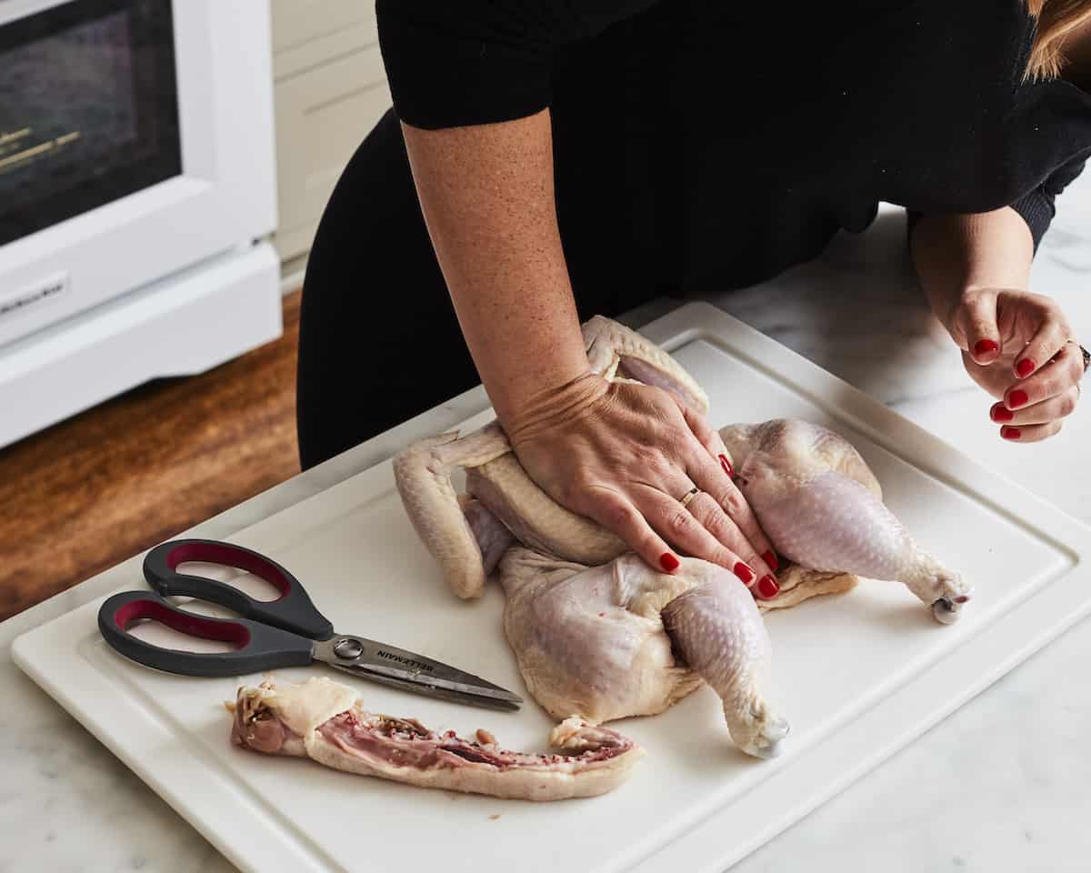 A woman smashing the backbone of a chicken to flatten it out, placed on a white cutting board, along with its cut out backbone and a pair of kitchen scissors.