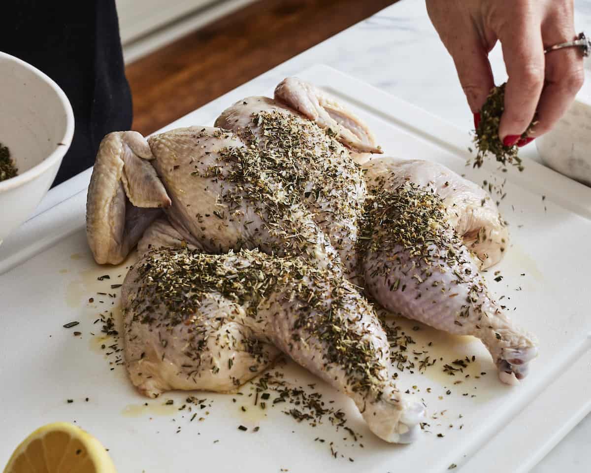 A woman sprinkling a mix of dried herbs onto a spatchcocked chicken placed on a white cutting board.