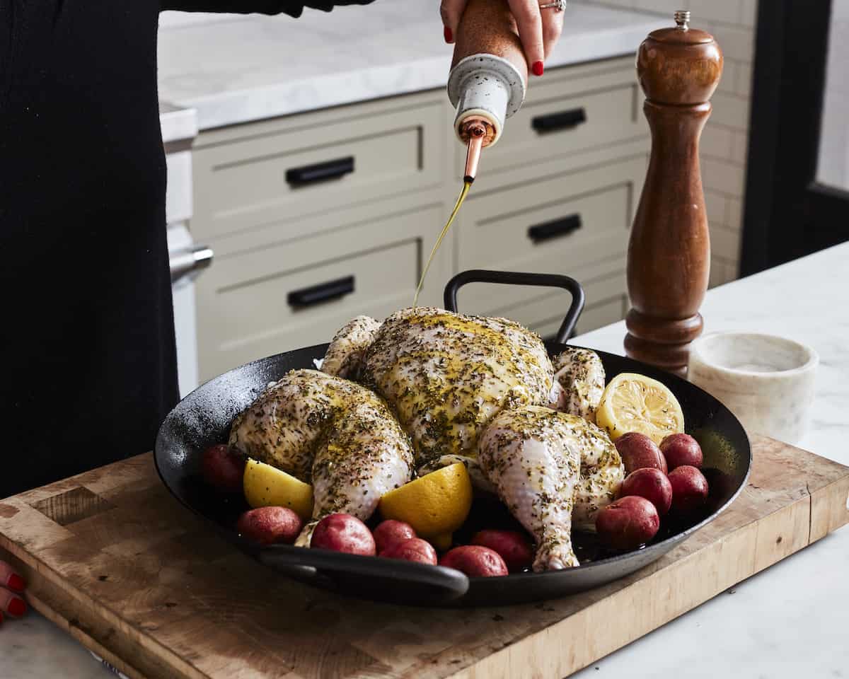A wooden cutting board with a skillet placed on it, containing lemon halves, baby potatoes and a spatchcocked chicken, and a woman drizzles olive oil onto it.
