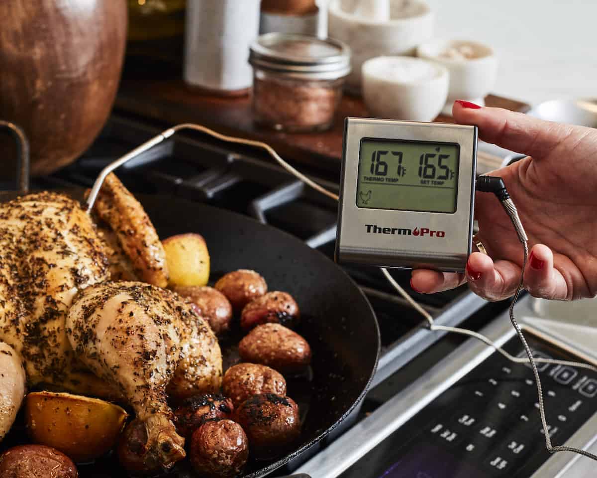 A woman holding a kitchen thermometer showing a temperature reading of 167F, and an expected temperature setting of 165F, with the thermometer probe in the breast of a roasted spatchcocked chicken in a skillet placed on a stovetop. 