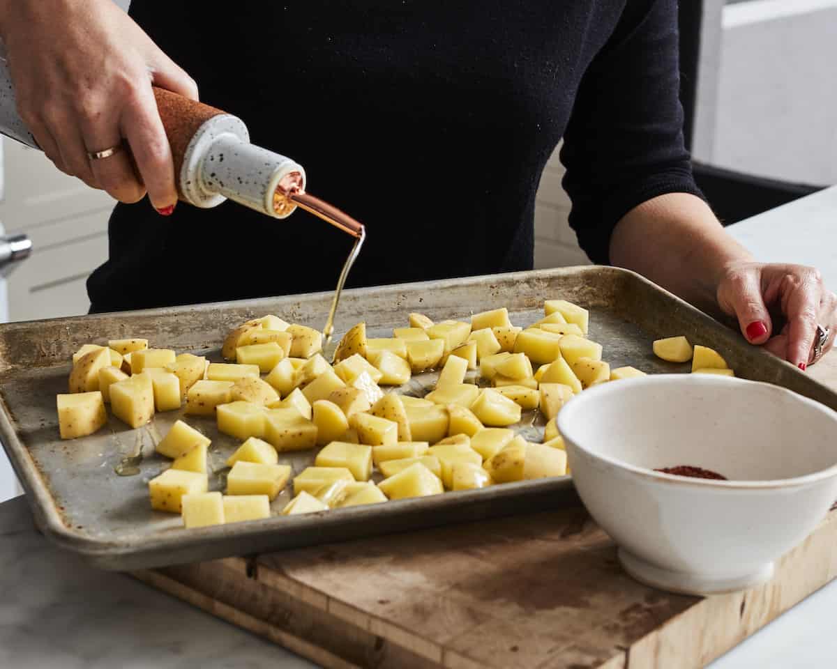 A woman pouring oil through a spout onto a baking sheet full of cubed potatoes with a small white bowl on the side with seasonings.