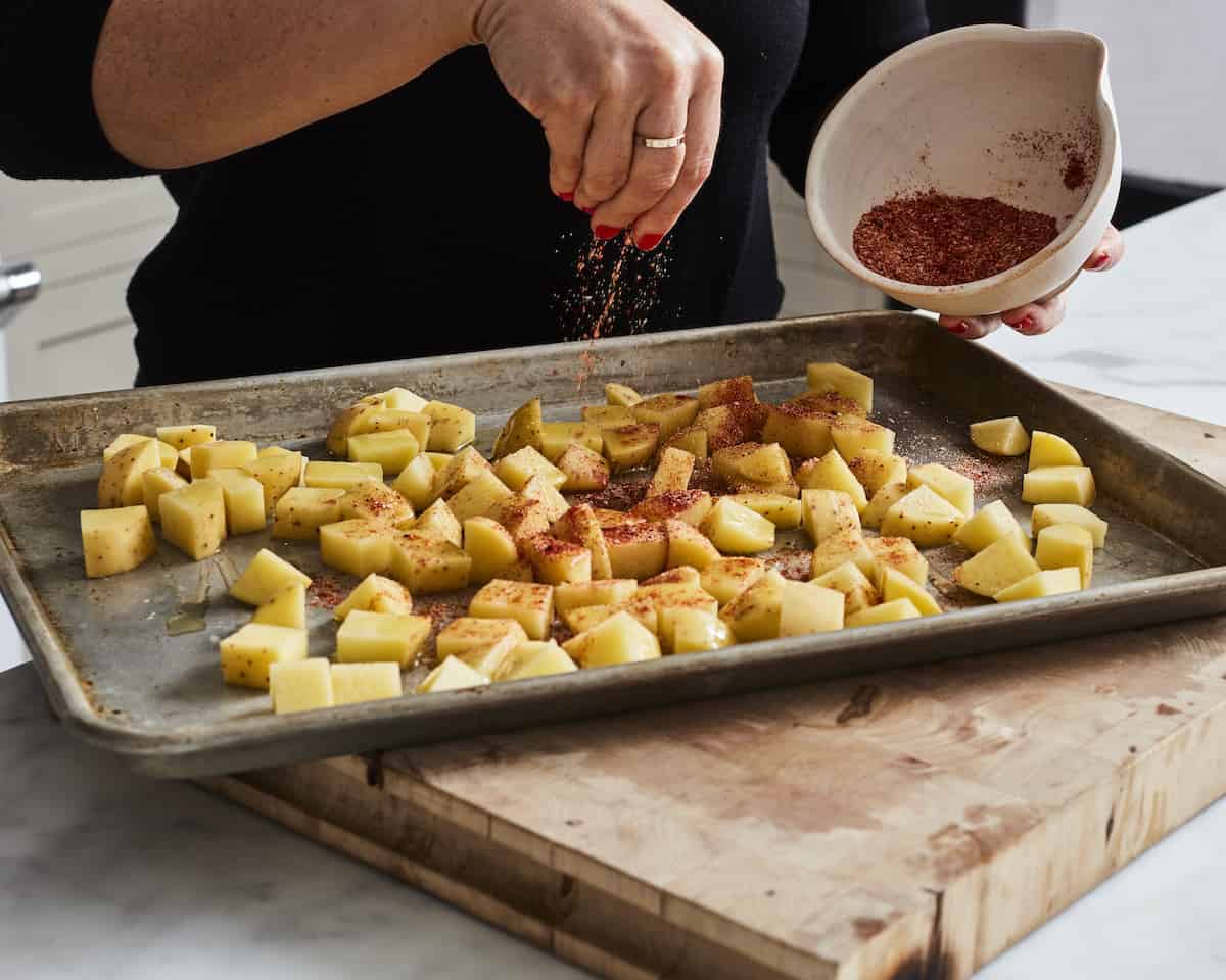 A woman sprinkling seasoning from a white bowl onto a baking sheet full of cubed potatoes.