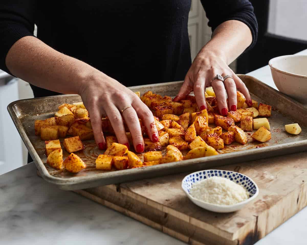 A woman tossing potatoes on a baking sheet full of cubed potatoes with a white and blue bowl of grated parmesan on the side.