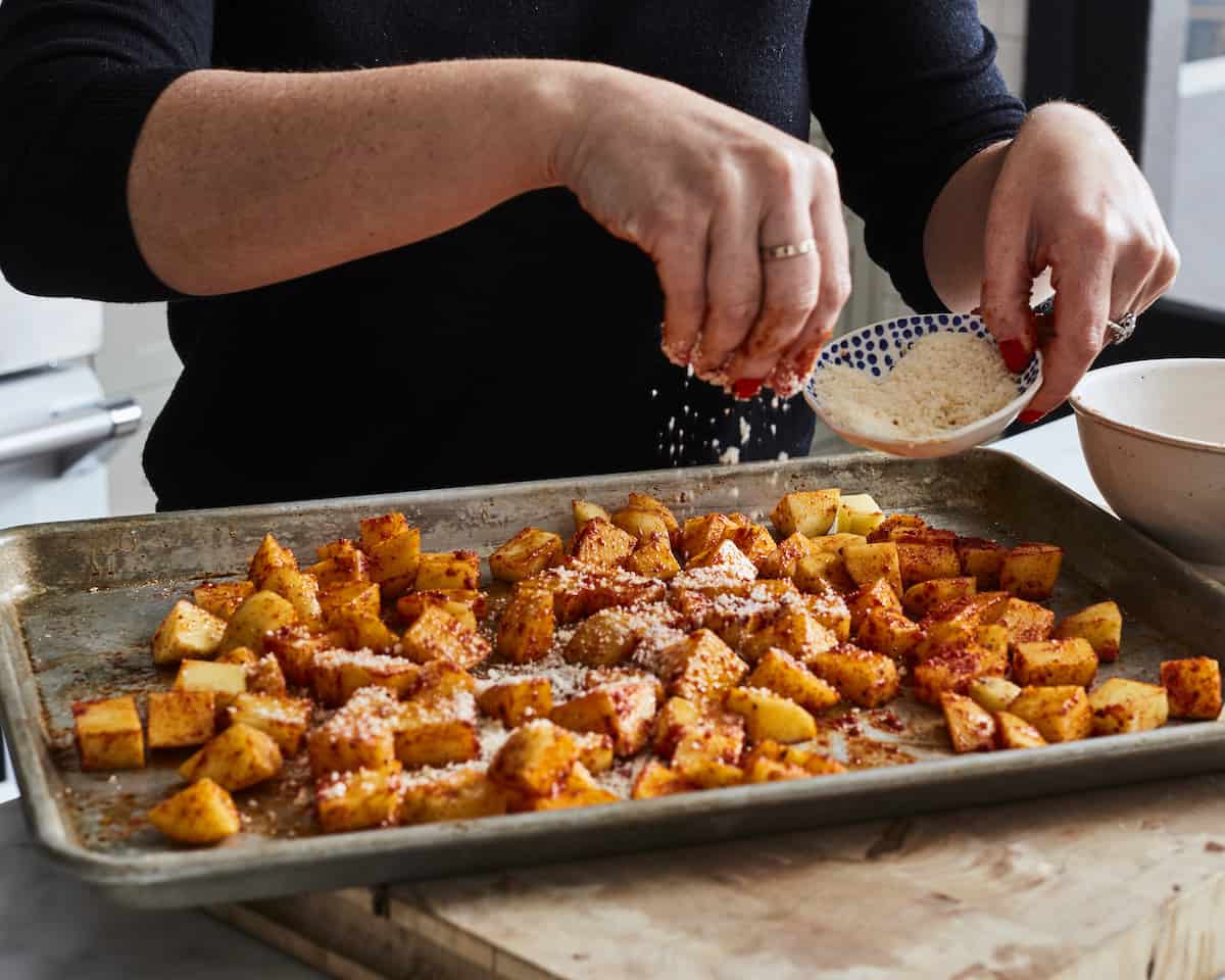 A woman sprinkling grated parmesan from a bowl onto a baking sheet full of cubed potatoes.