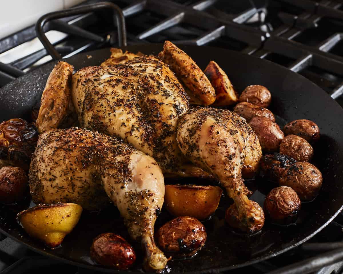 A skillet closeup, with a roasted spatchcock chicken on a bed of lemon halves and baby potatoes on a stovetop.