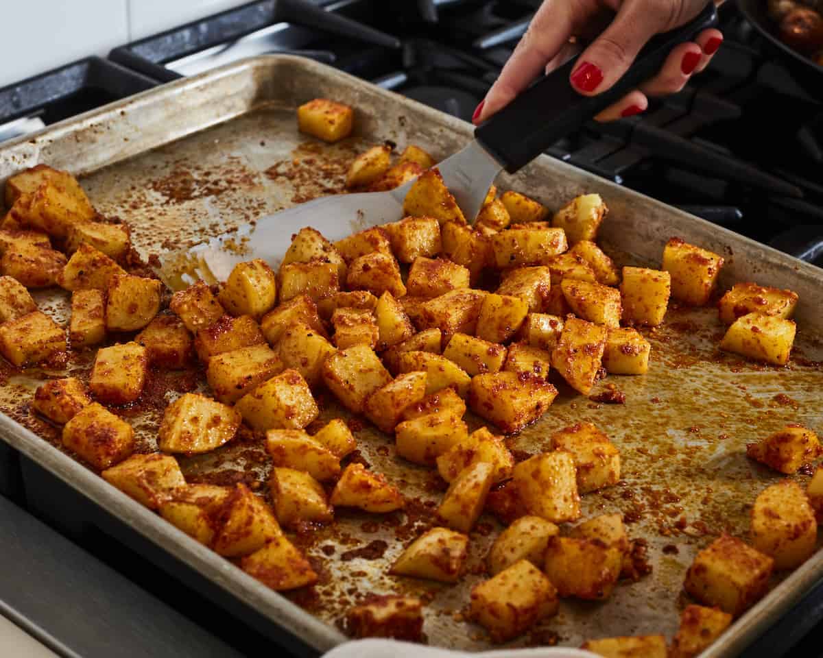 A woman tossing cubed partially roasted potatoes on a baking sheet with a flat spatula.