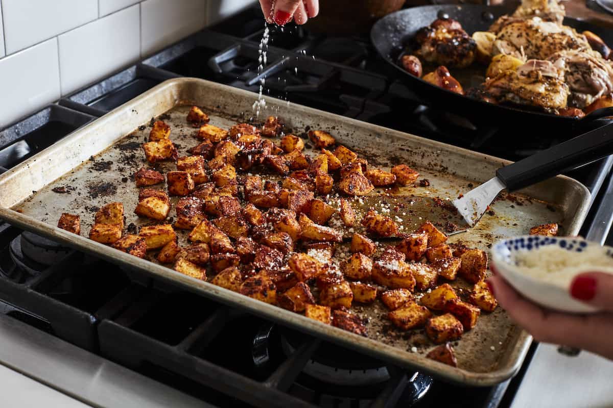 A woman holding a little bowl of grated parmesan, sprinkling it into a baking sheet full of roasted potatoes.
