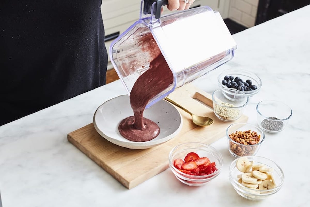 Gaby pouring the blended acai mix into a shallow white bowl, with little bowls of swtrawberries, banana, blueberries, granola and toasted coconut on the side.