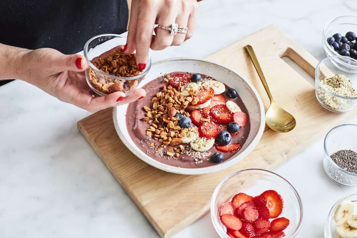 Gaby topping an acai bowl with granola. The shallow white bowl of acai is already topped with sliced strawberries, banana, blueberries with extra toppings in little bowls on the side.