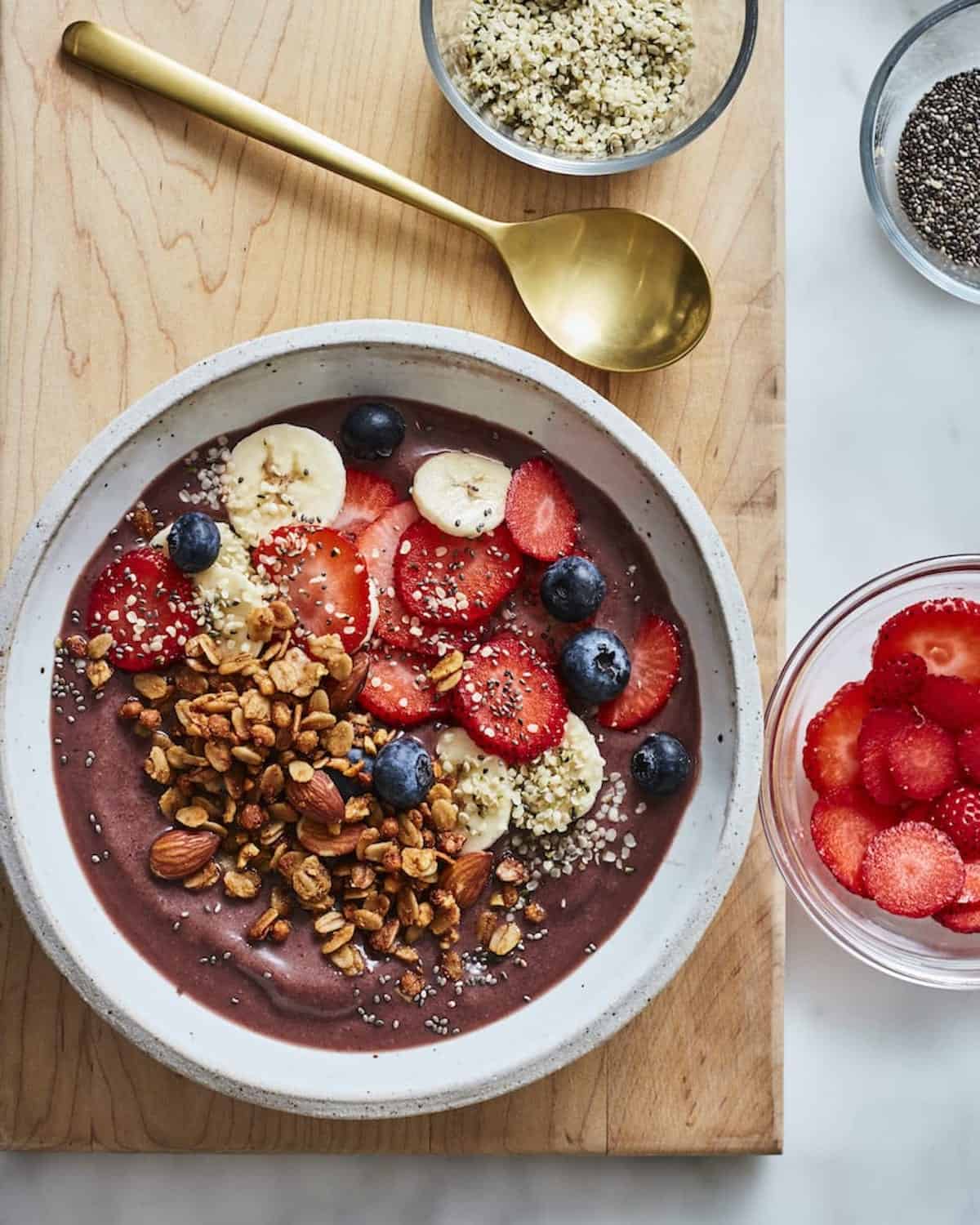 A shallow white acai bowl topped with  sliced strawberries, banana and blueberries, and granola, with a small bowl of sliced strawberries and a golden spoon on the side.