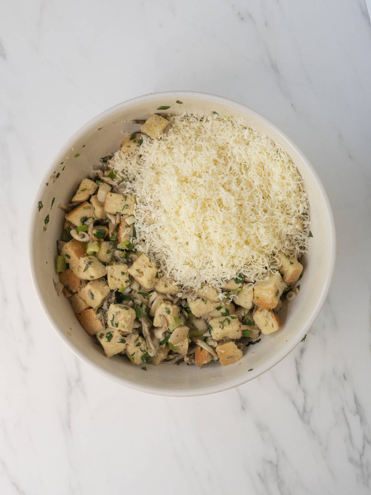 A white mixing bowl with cooked mushrooms, dried bread cubes, eggs and broth mix, and grated parmesan.