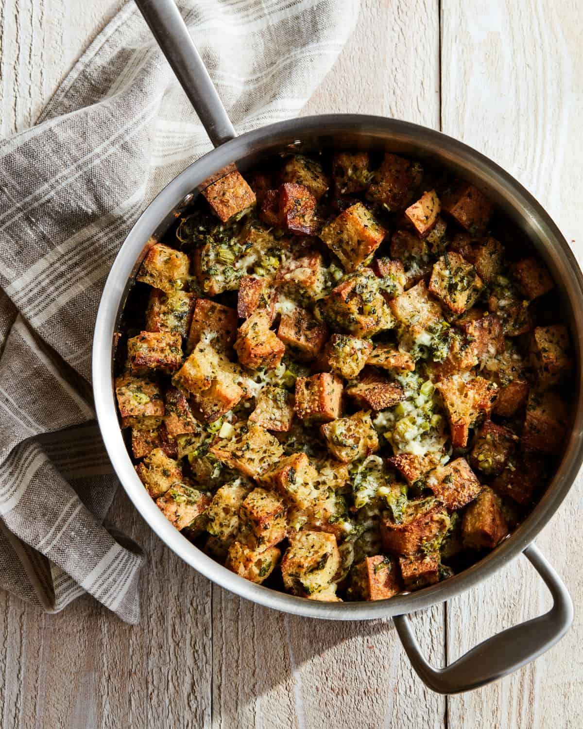 A steel skillet with rustic herb stuffing, baked and browned on top, placed on a grey and white kitchen towel.