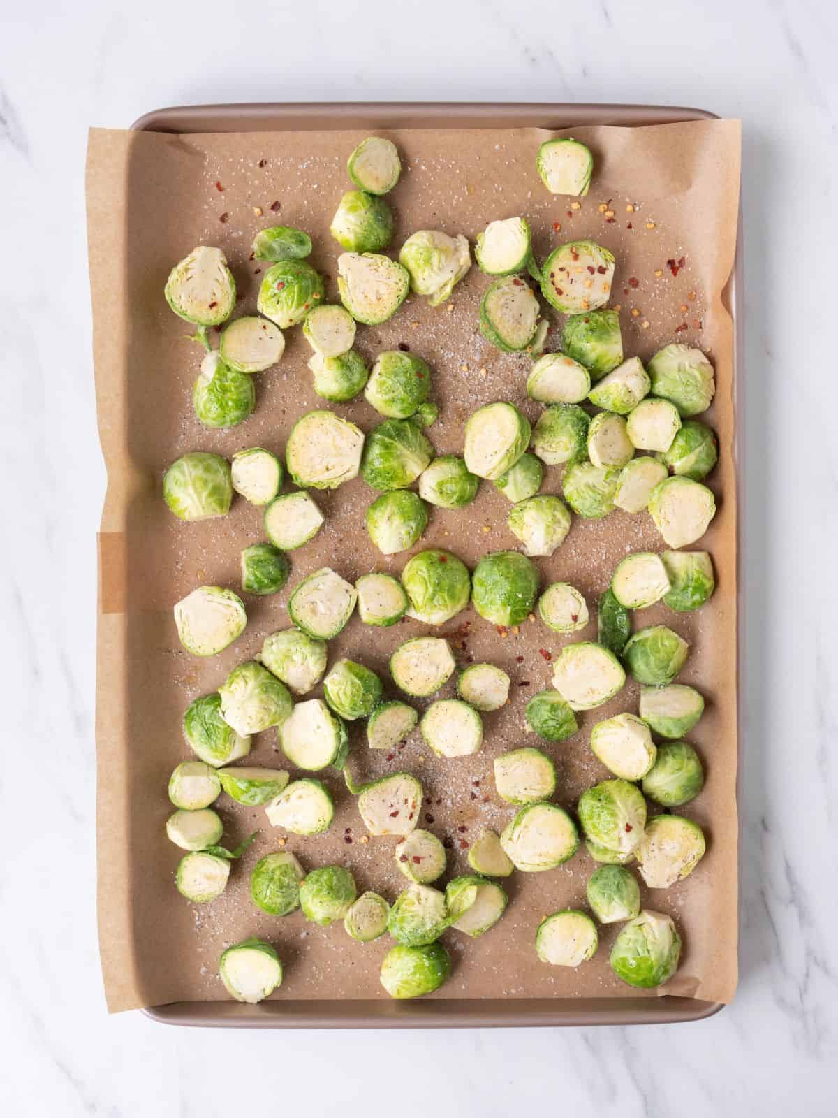 A baking sheet pan lined with parchment paper with halved raw brussels sprouts laid out, coated in oil, salt, pepper and red pepper flakes.