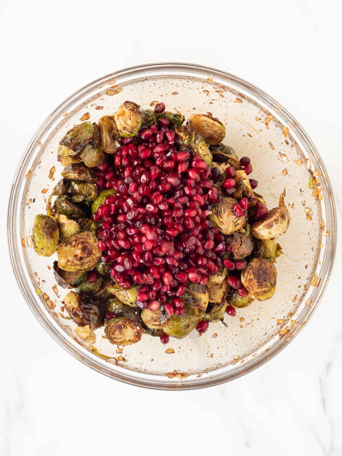 A glass mixing bowl with roasted brussels sprouts salad topped with pomegranate seeds.