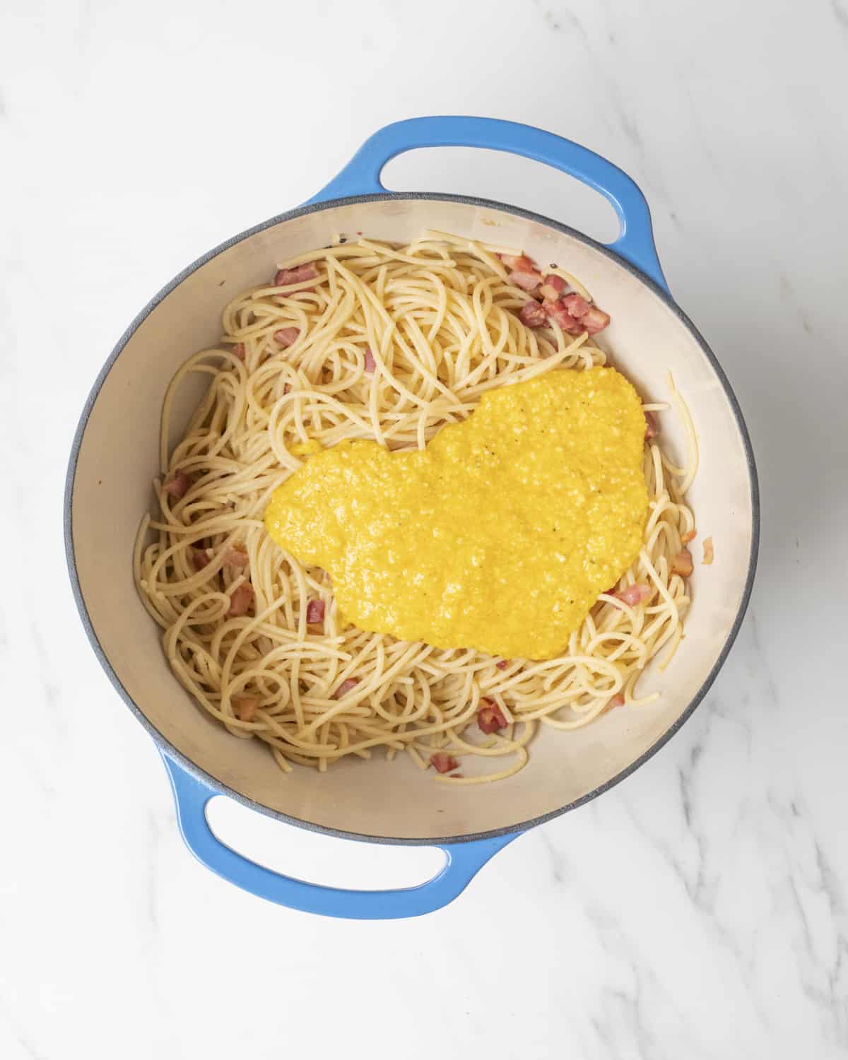 A blue Dutch oven skillet with cooked spaghetti noodles and pancetta topped with the egg mixture.