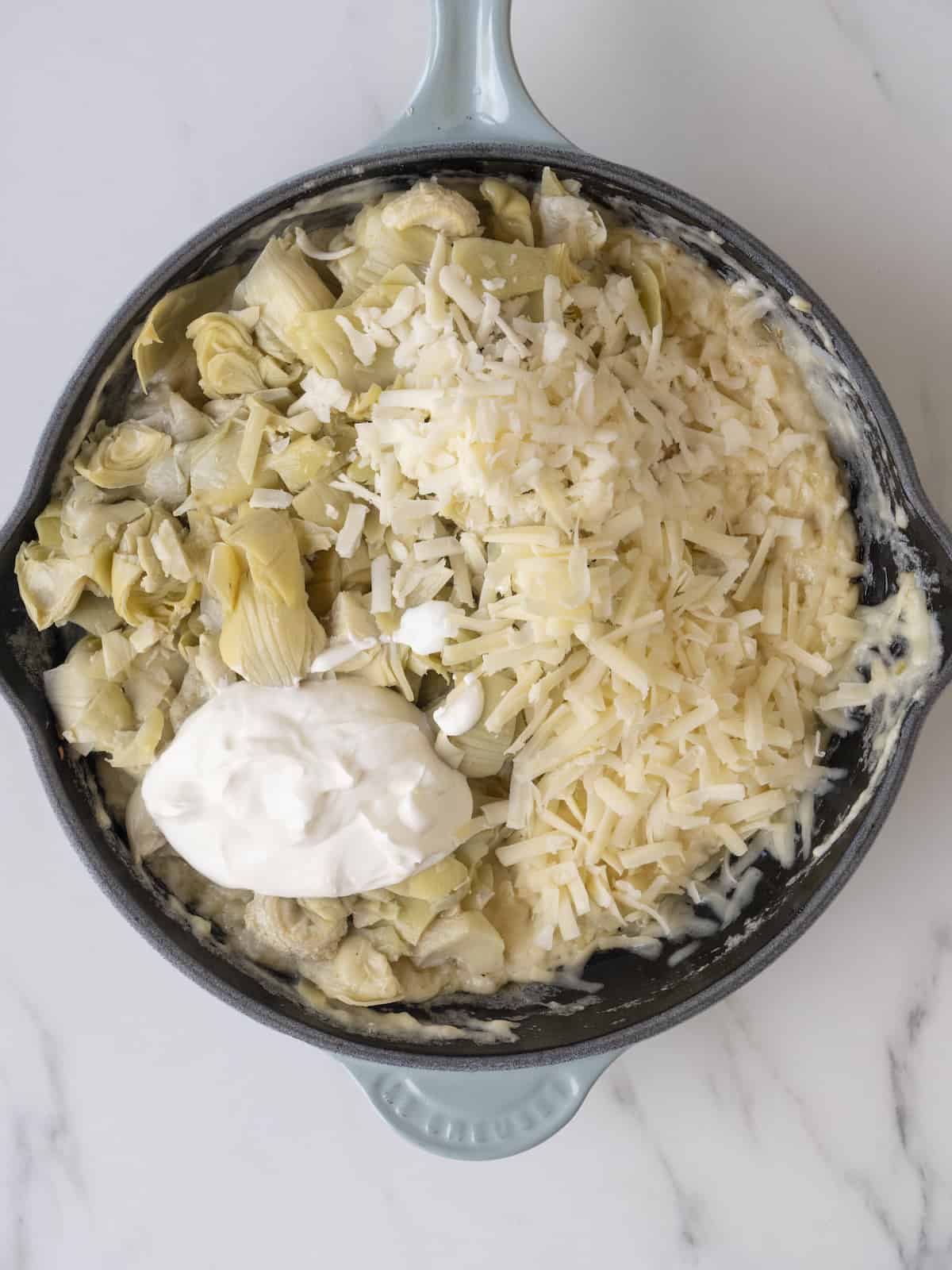 A skillet with grated cheeses, artichoke hearts and sour cream just added.