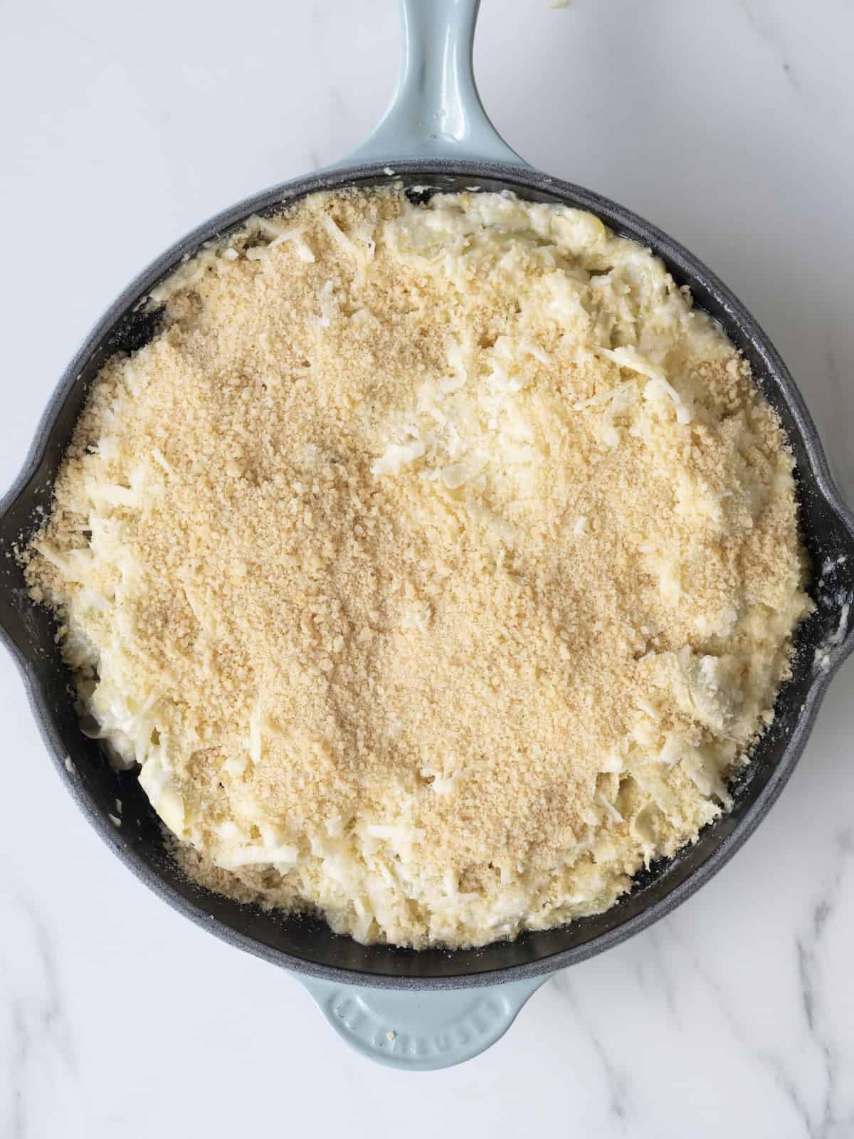 A skillet with cheesy artichoke dip topped with a mixture of parmesan, gruyère, fontina and panko breadcrumbs, ready to be placed in an oven for baking.