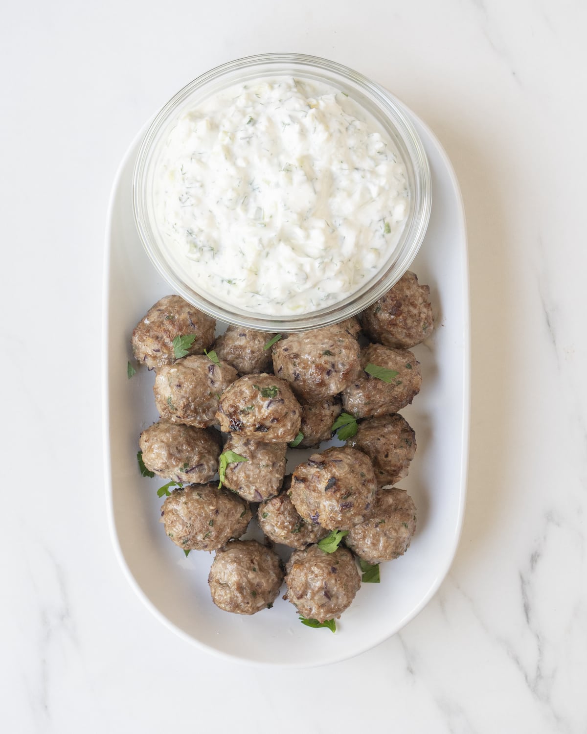 Greek lamb meatballs topped with parsley leaves on a white serving tray beside a clear bowl of tzatziki sauce.
