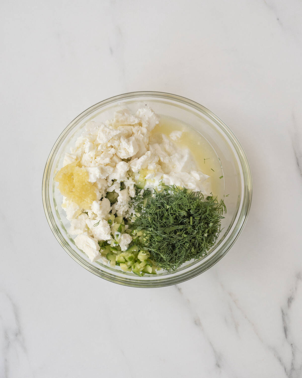 A clear bowl of greek yogurt, lemon juice, parsley, dill, cucumber, and garlic.