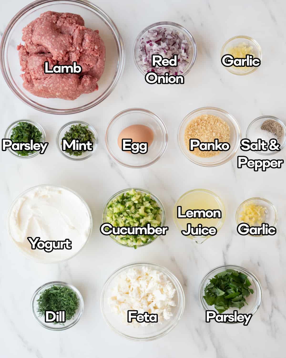 Ingredient shot of individual ingredients in clear bowls including lamb, red onion, garlic, parsley, mint, egg, panko breadcrumbs, salt & pepper, yogurt, cucumber, lemon juice, dill, and feta 