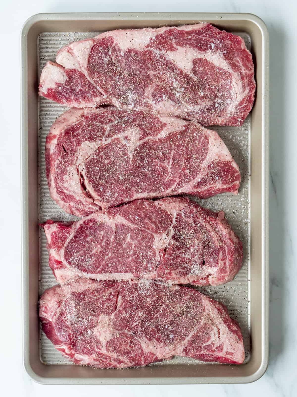 A baking sheet with ribeyes seasoned with lots of salt and pepper.