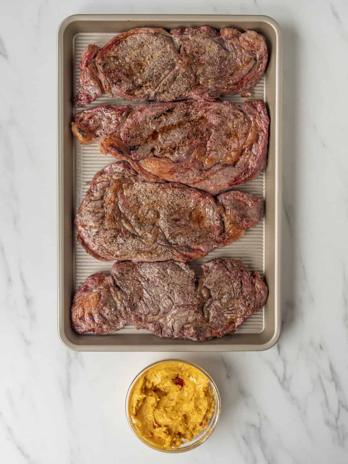 A baking sheet with grilled ribeyes seasoned and a small bowl of calabrian chile compound butter on the side.