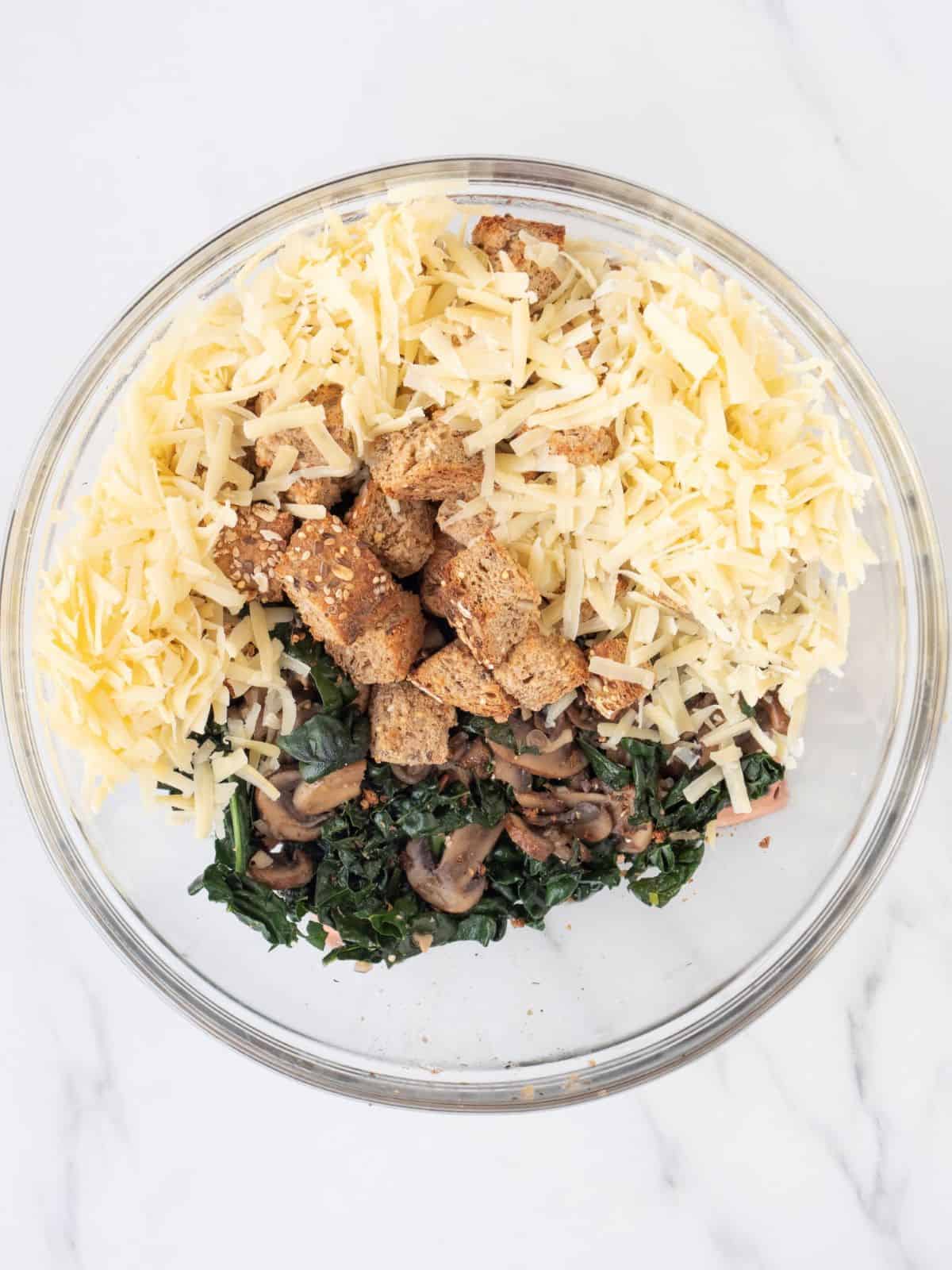 A glass mixing with with the mushroom and kale mixture that was cooked, along with shredded cheese and bread cubes. 