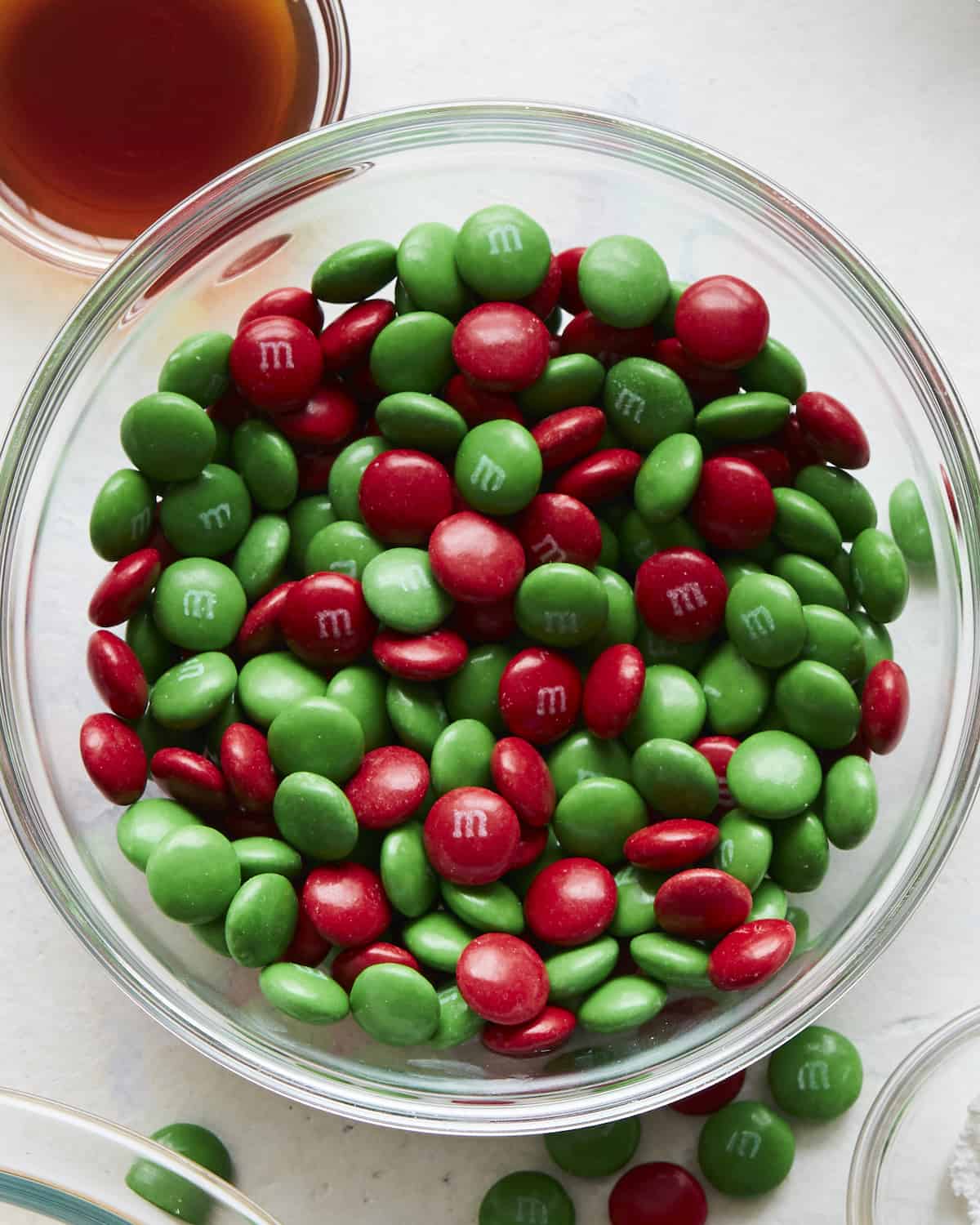 A glass bowl with red and green M&Ms with some M&Ms outside the bowl on the counter.