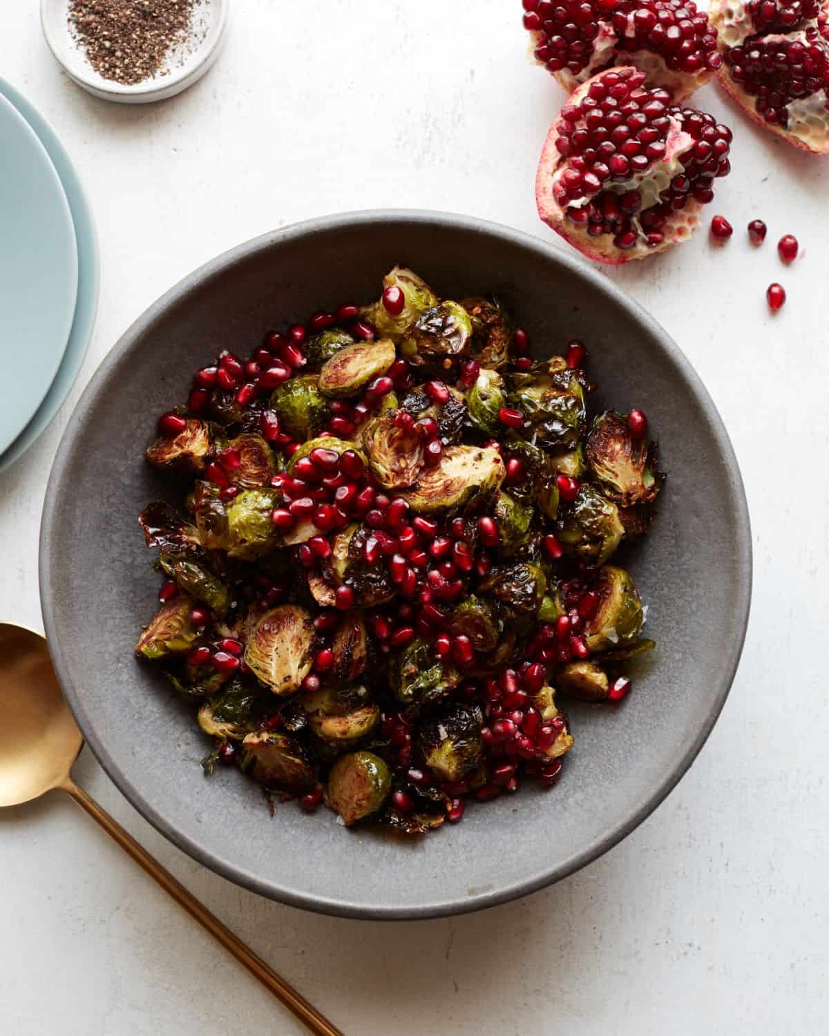 A grey shallow bowl with roasted brussels sprouts salad topped with pomegranate seeds, with a gold spoon on its side, pomegranate quarters and plates next to it.