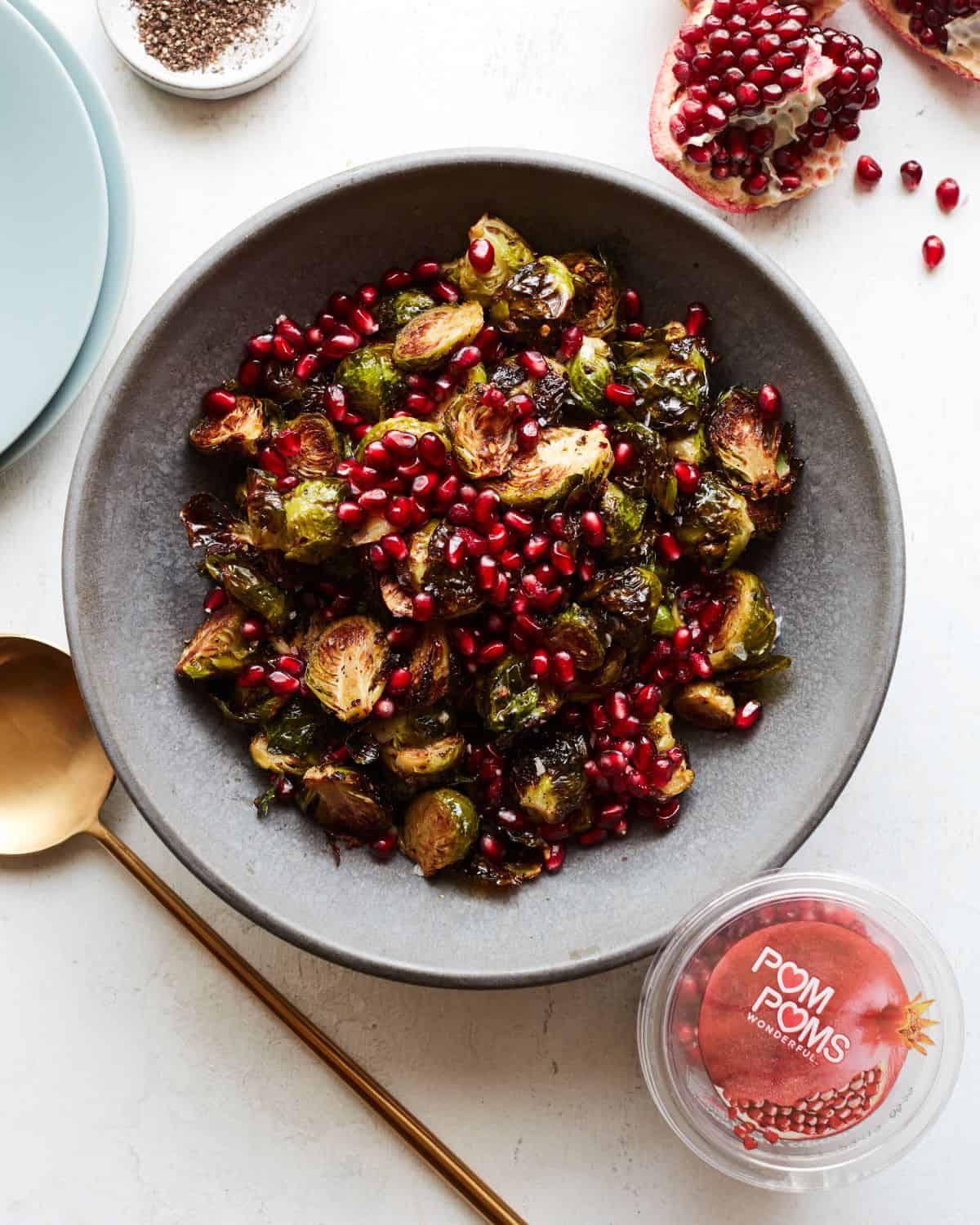A grey shallow bowl with roasted brussels sprouts salad topped with pomegranate seeds, with a gold spoon on its side, pomegranate quarters, plates and a box of POM POMS full of pomegranate seeds next to it.
