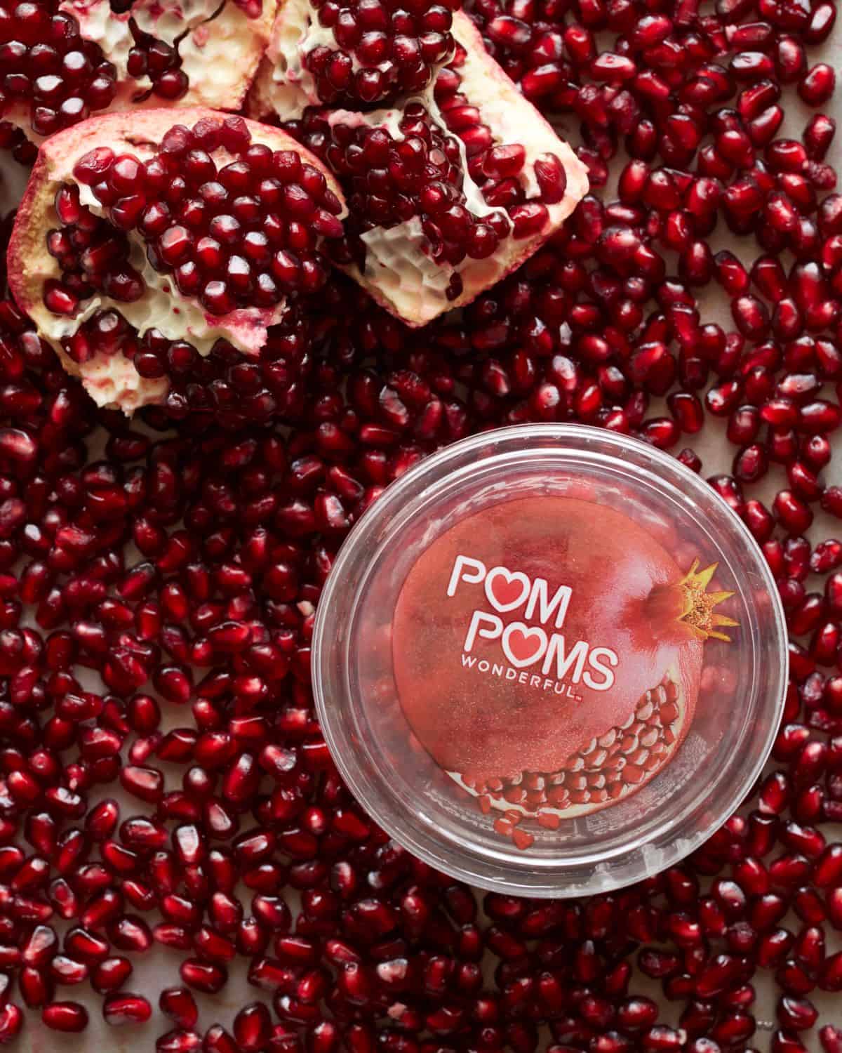 A close up shot of POM POMS box of pomegranate seeds placed on a counter full of pomegranate seeds and pomegranate quarters.