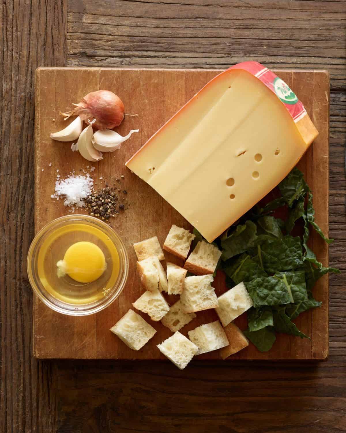 A wooden board with salt, pepper, garlic cloves, shallot, bread cubes, chopped kale, a small glass mixing bowl with a broken egg and a big block of Parrano cheese.
