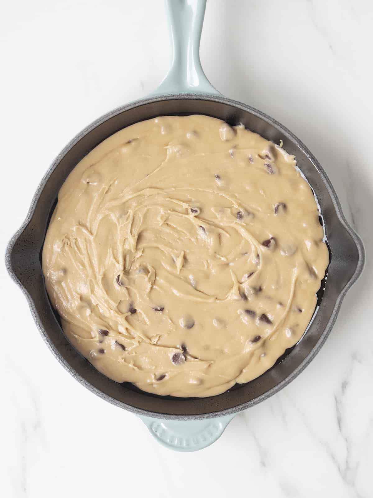 A skillet with a blue handle, with cookie dough spread out evenly in the skillet.