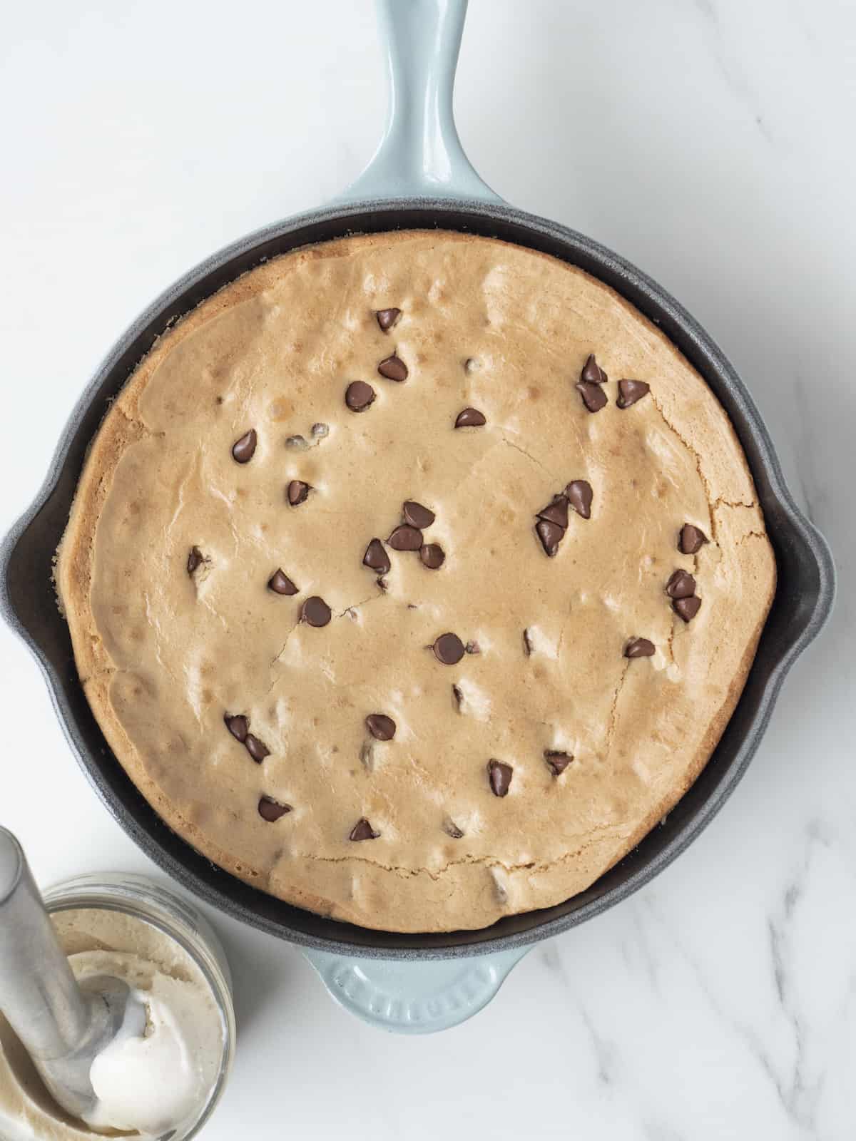 A skillet with a blue handle, with a giant chocolate chip cookie, called, pizookie, baked in it, with a pint of vanilla ice cream on the side with an ice cream scooper in it.