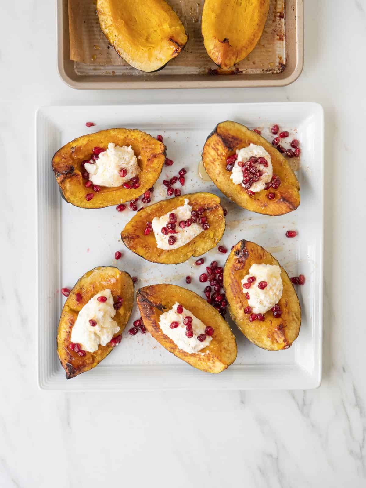 A white square plate with roasted acorn squash topped with ricotta and pomegranate seeds and drizzled with honey and nutmeg, with a partially visible baking sheet with roasted acorn squash.