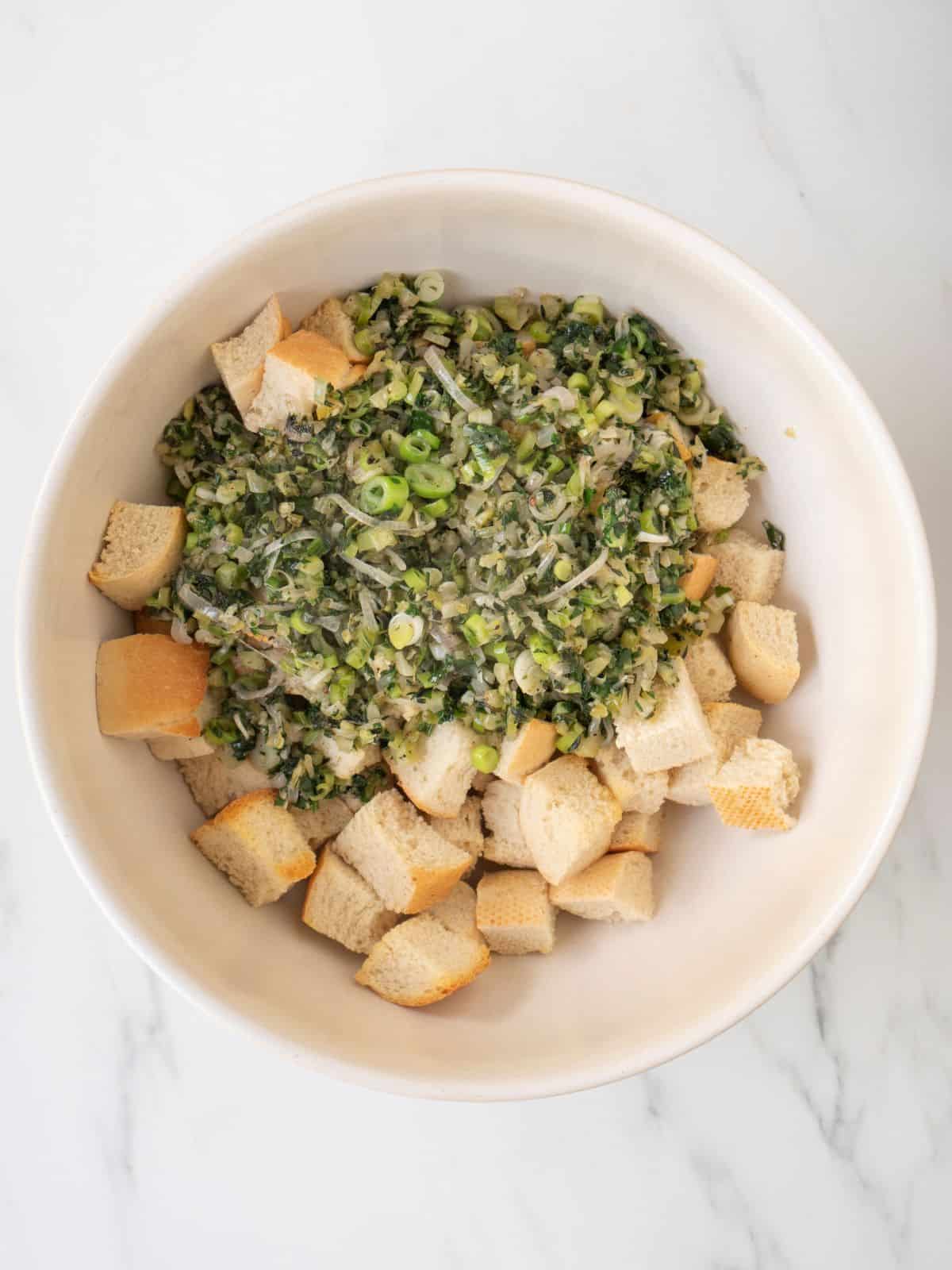 A white mixing bowl with cubed bread, and the cooked herb and vegetable mixture.