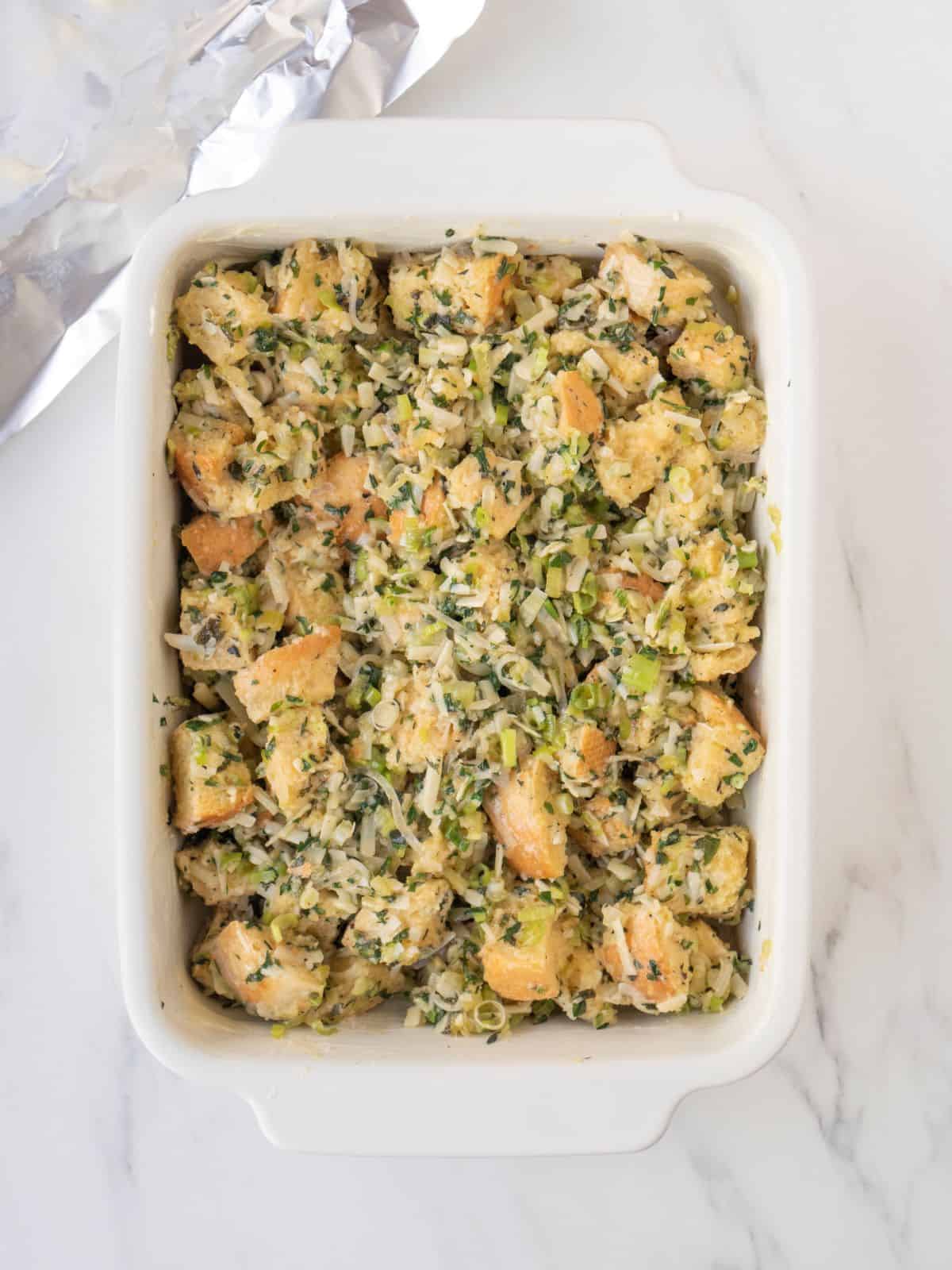 A white rectangular baking dish with rustic herb stuffing read to be baked, with a piece of foil on the side.